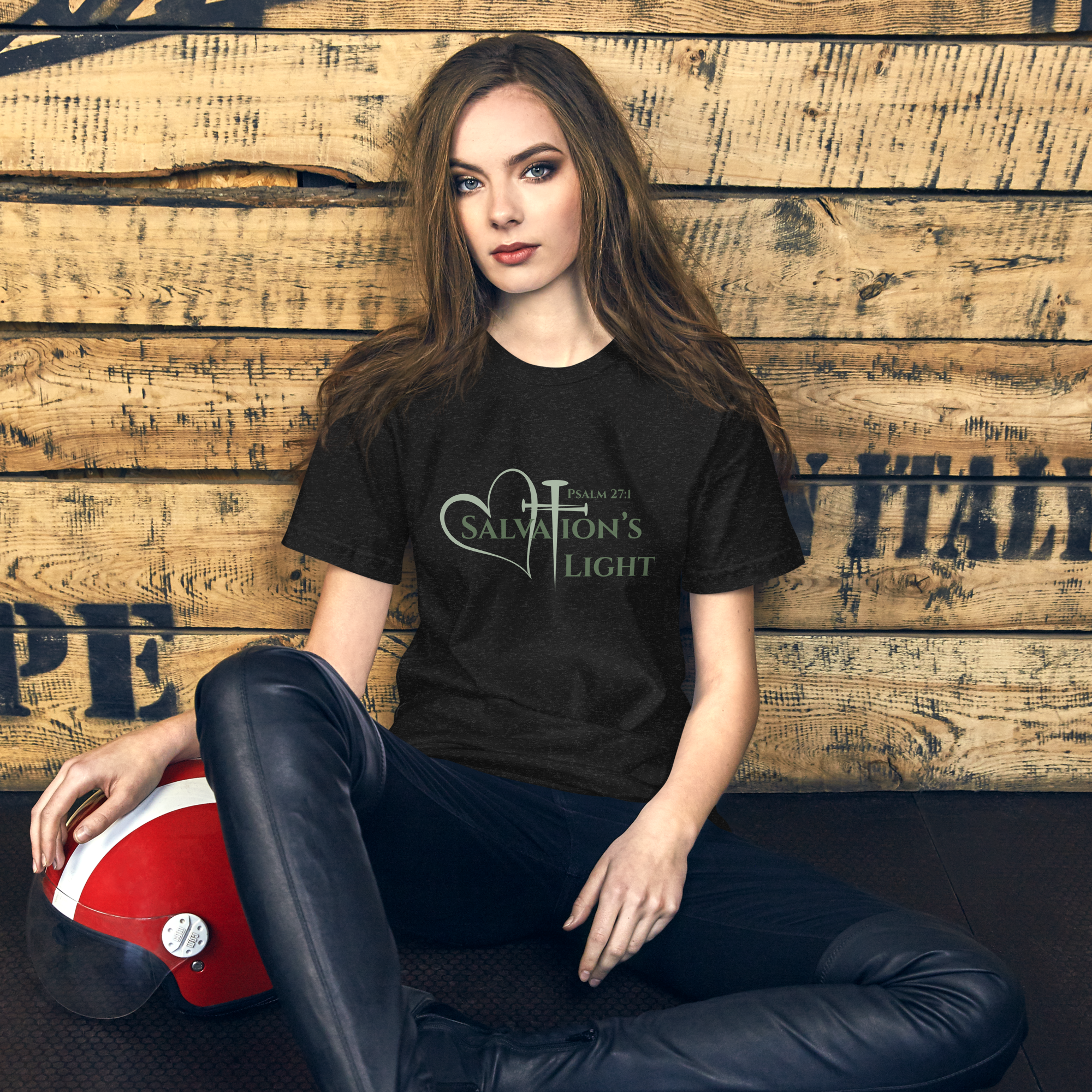 Female model sitting against a wall wearing a short sleeve t-shirt from the Salvation’s Light Signature Collection featuring Psalm 27:1 and the words “Salvation’s Light,” with the letter T designed as a sage green three-nail cross and a matching sage green heart