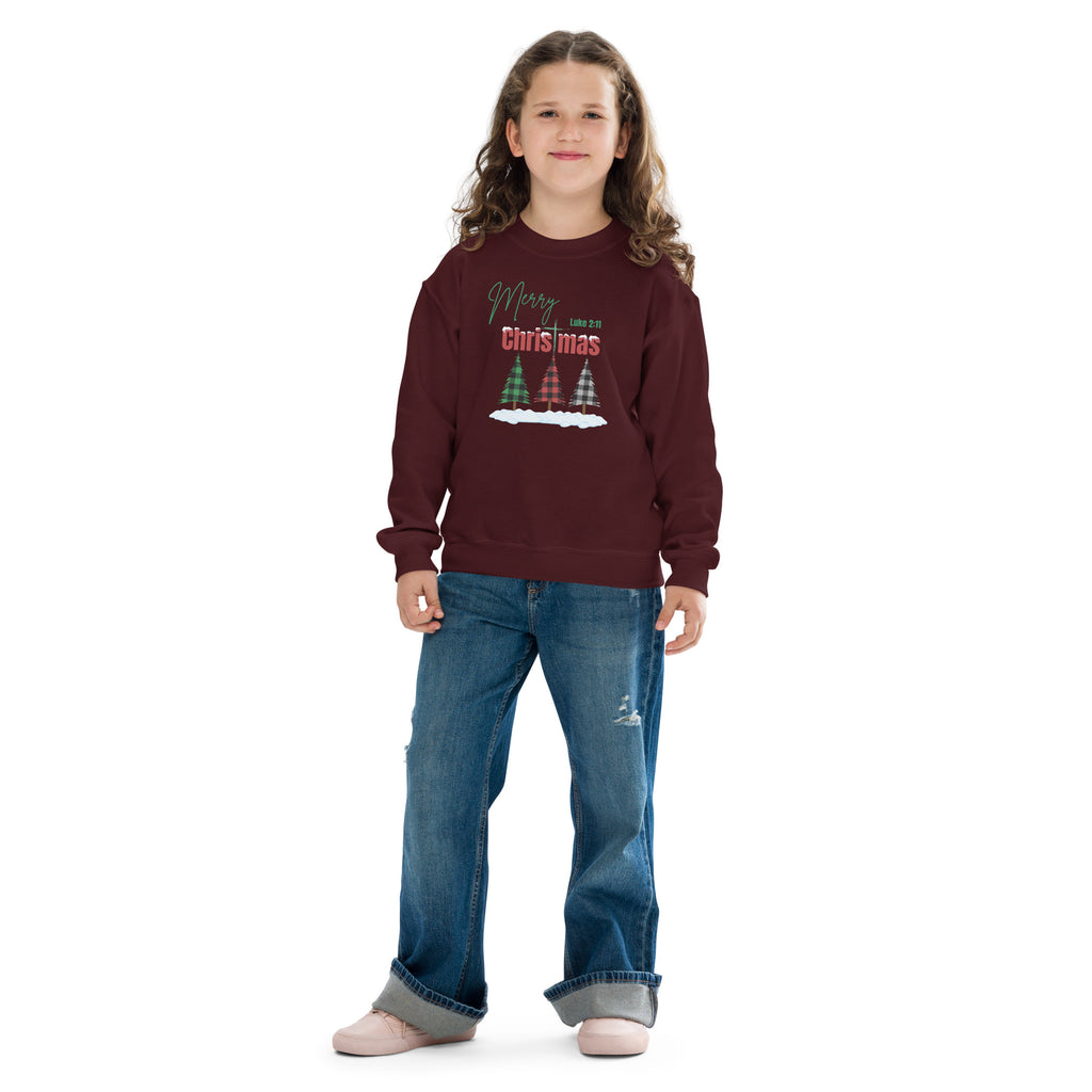 Girl wearing maroon youth Christmas sweatshirt featuring plaid Christmas trees, snow on the ground, Merry Christmas wording, Luke 2:11, and cross-shaped T