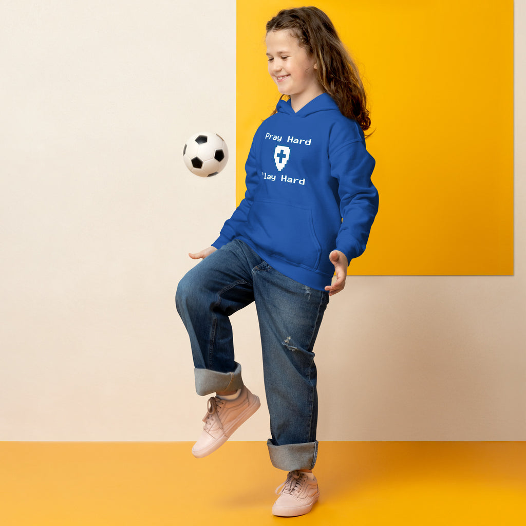 Girl bouncing a soccer ball on her knee wearing a kids Christian hoodie in royal blue showing the front design text “Pray Hard” above a shield with a cross and the text “Play Hard” below in a gamer pixel font