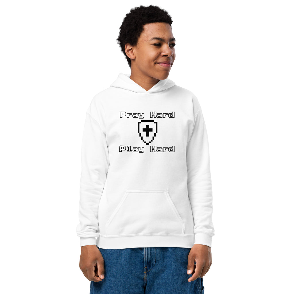 Boy wearing a kids Christian hoodie in white showing the front design text “Pray Hard” above a shield with a cross and the text “Play Hard” below in a gamer pixel font