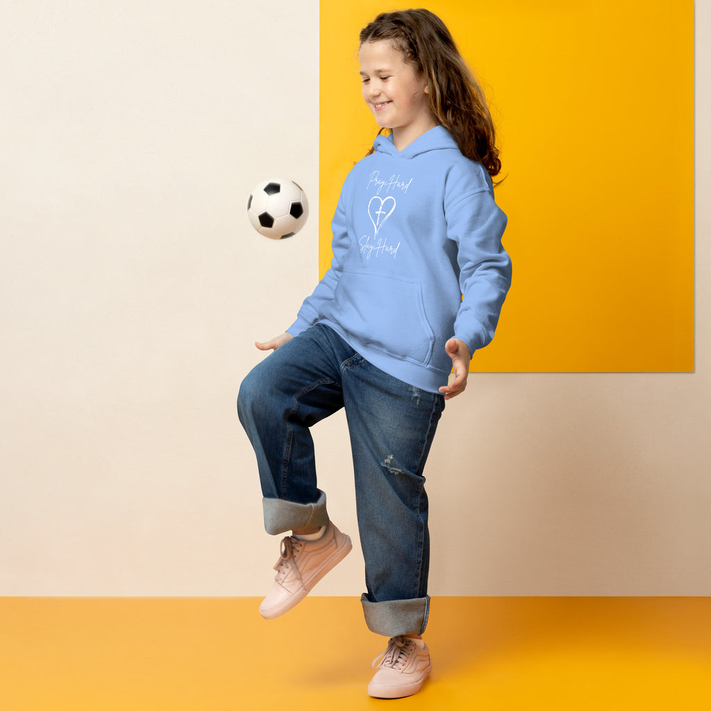 Girl model bouncing a soccer ball on her knee while wearing a carolina blue girls Christian hoodie featuring the exact text “Pray Hard” and “Slay Hard” in script font with a heart and a cross in the center between the phrases