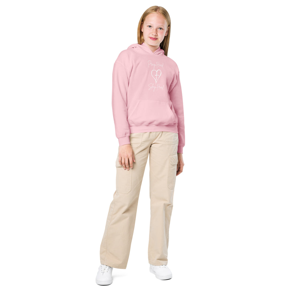 Full body image of a girl wearing a light pink girls Christian hoodie featuring the exact text “Pray Hard” and “Slay Hard” in script font with a heart and a cross in the center between the phrases