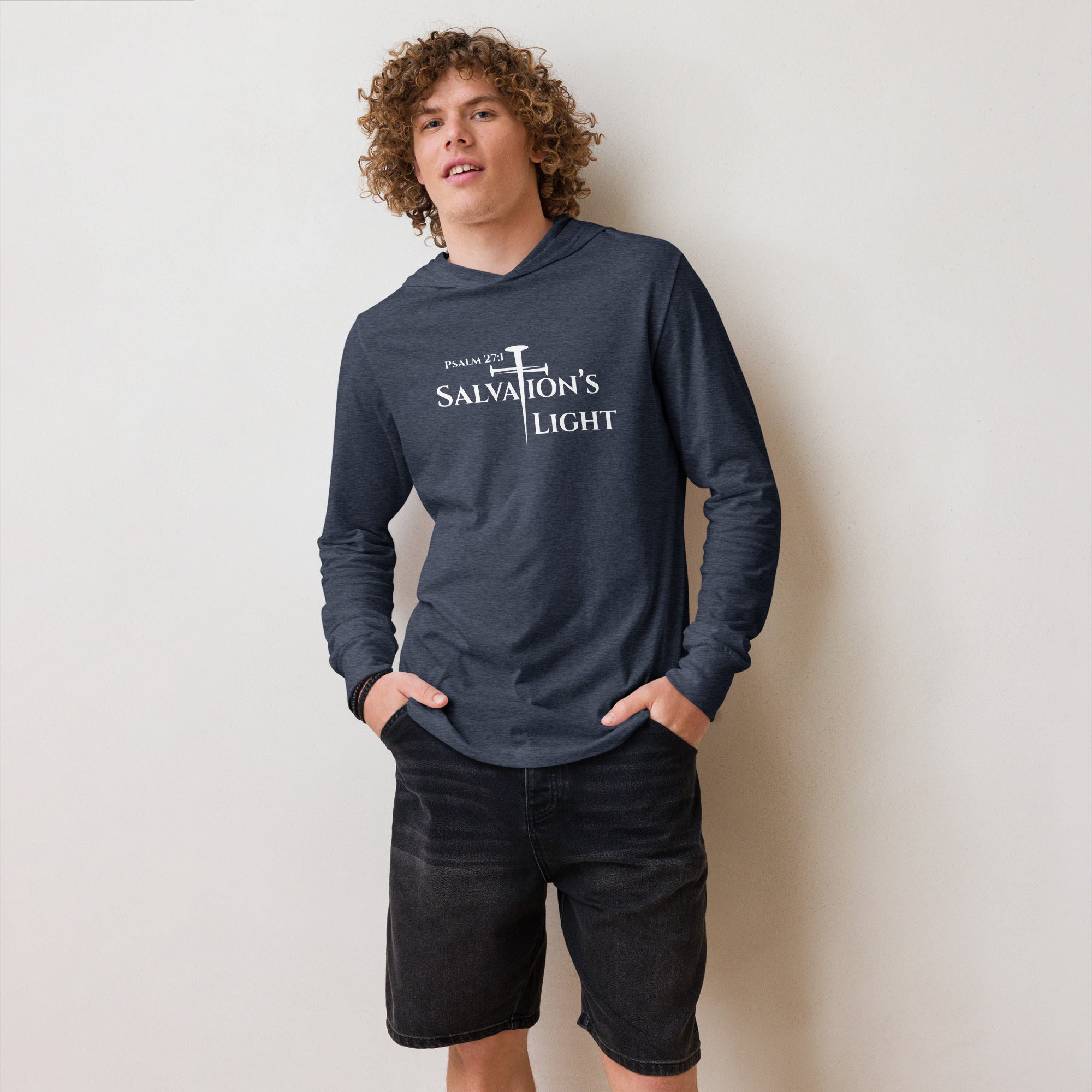 Front view of a male model standing against a tan wall wearing the heather navy men’s Christian hooded long sleeve tee featuring the Salvation’s Light design with the three-nail cross forming the “T” in Salvation and the text “Psalm 27:1.”