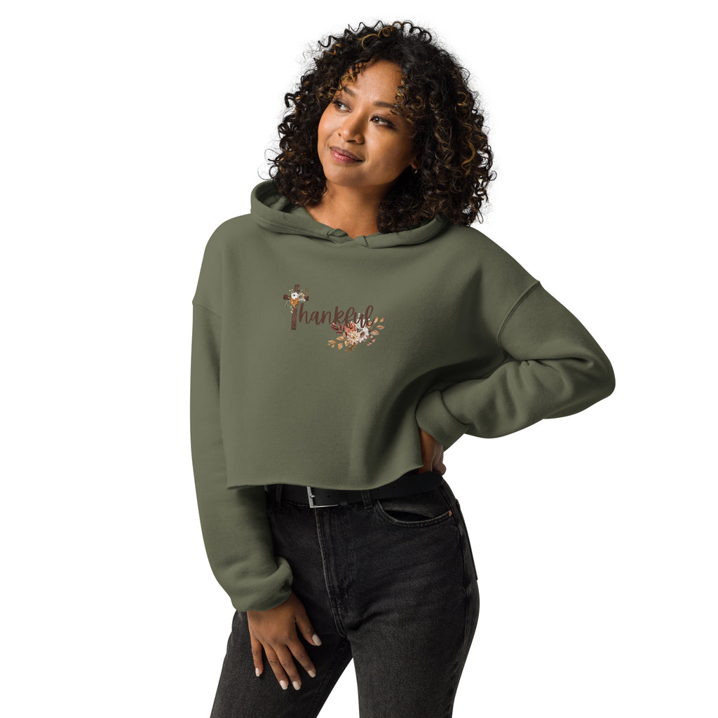 Front view of a female model wearing a women’s cropped Christian hoodie in military green featuring the word “Thankful” in a brown script font, the “T” designed as a wooden cross, and fall floral accents.