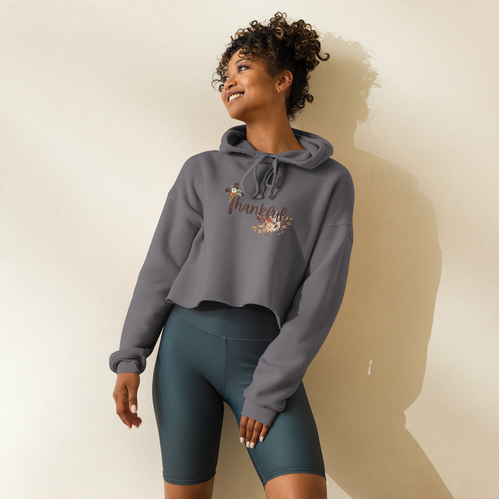 Front view of a female model wearing a women’s cropped Christian hoodie in storm displaying the “Thankful” brown script text with a wooden cross forming the “T” and soft autumn floral accents.