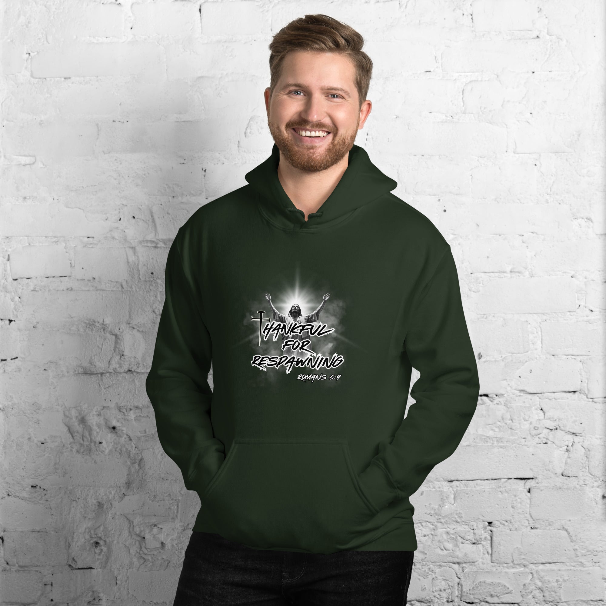 Male model against a white brick wall wearing a black Christian gamer humor hoodie featuring “Thankful for Respawning,” Romans 6:9, a resurrection image of Jesus, and a cross formed by three nails