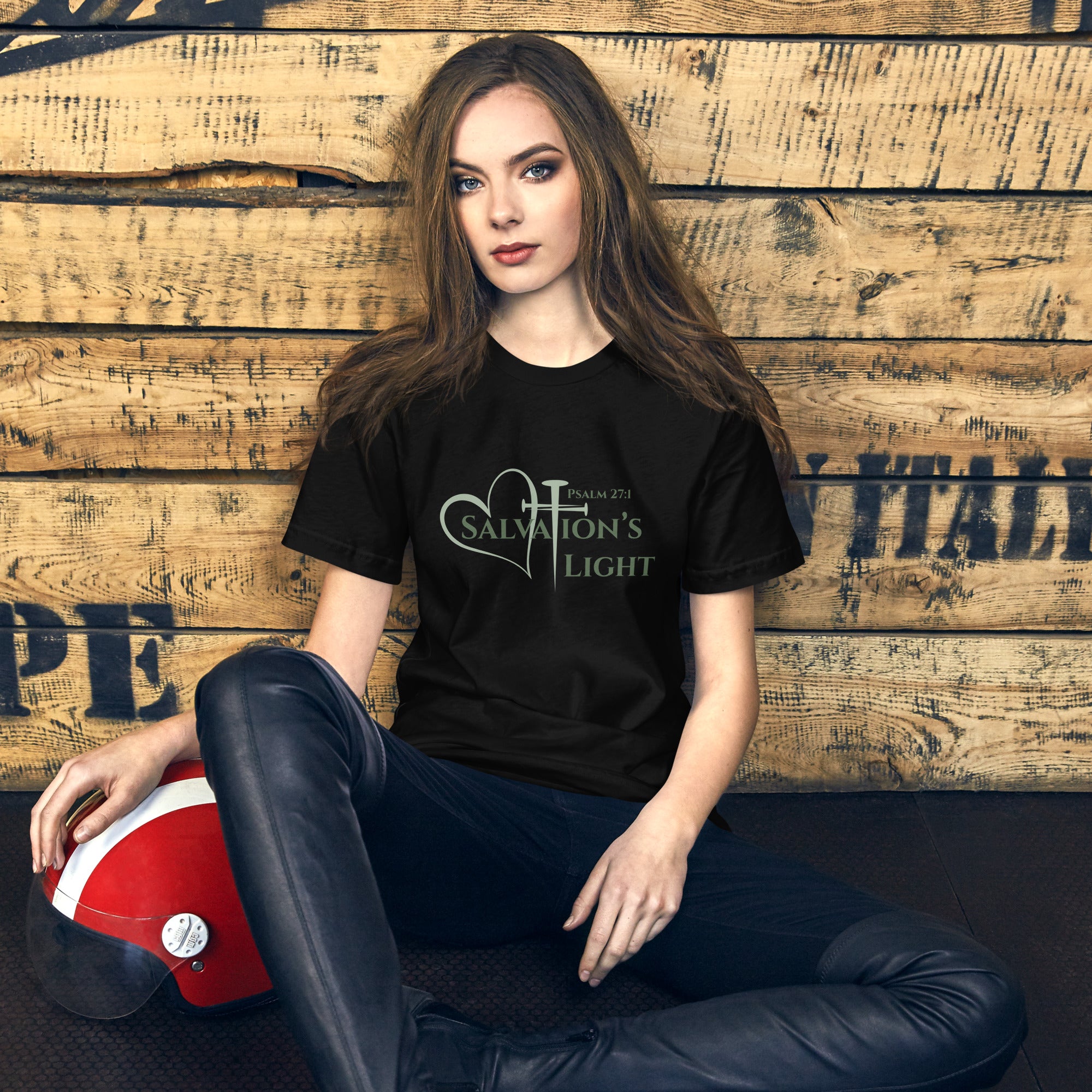 Female model wearing a black short sleeve t-shirt from the Salvation’s Light Signature Collection, featuring Psalm 27:1 and the word “Salvation’s Light,” with the T designed as a sage green three-nail cross and accompanied by a sage green heart